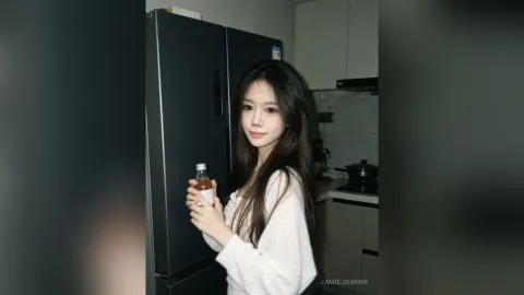 JamieJoanne — JamieJoanne stands in a kitchen wearing a white long-sleeved shirt while holding a small glass bottle.