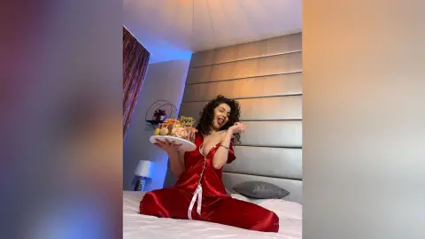 Cheerful woman in red satin pajamas holding a dessert platter in a modern bedroom setting.