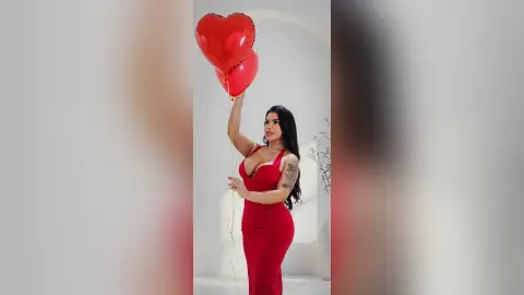 Glamorous woman in a red evening dress holding red heart balloons in a bright studio setting.