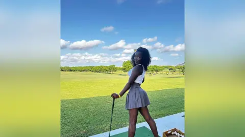 Young woman in a white tank top and grey skirt standing on a sunny green golf course field.