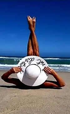 Relaxed person lying on a sunny beach with legs up, wearing a white sun hat against a blue ocean background.