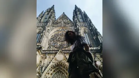 Young woman in casual attire standing before the majestic, historic St. Vitus Cathedral under a cloudy sky.