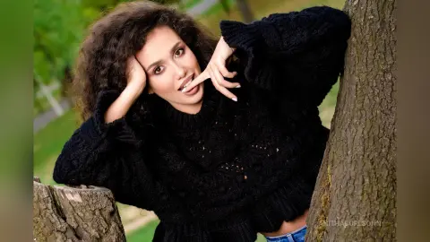 Smiling woman with curly hair in a cozy black knit sweater leaning against a tree in a lush green park.