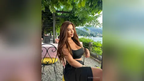 ReynaGomes poses outdoors in a black crop top and skirt, sitting at a table under green leaves with a lake and mountains in the background.