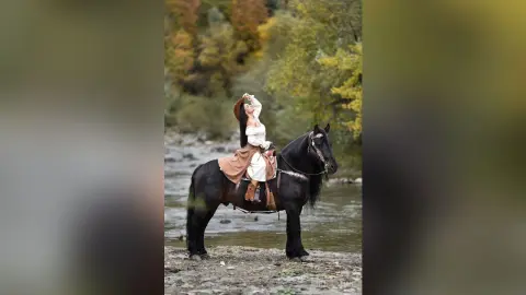 Woman in white bohemian dress riding a black horse through a scenic river in a lush autumn forest.