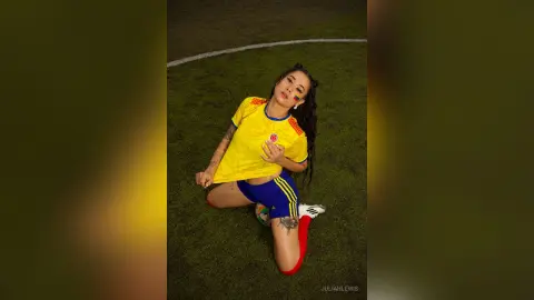 JuliahLewis kneels on a grass field wearing a yellow and blue soccer jersey and matching shorts.