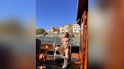 LucyMartin sits on a metal bench on a boat, wearing a white bra and tan trousers against a backdrop of a river and city buildings.