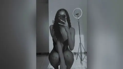 Young woman in a black bodysuit and fishnet tights taking a moody selfie in front of a ring light.