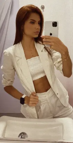AprilJakovish poses in a white blazer and matching crop top and pants, showing her midriff and long brown hair in a mirror selfie.