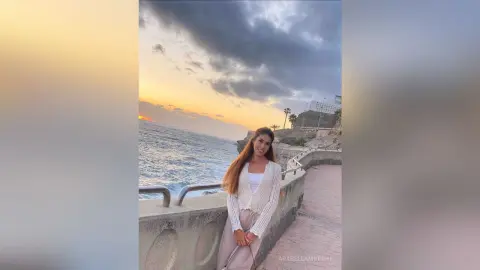 Woman in a white knit cardigan standing by the ocean at sunset in a dreamy coastal setting.