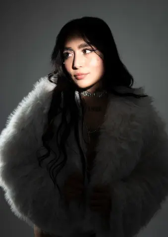 Young woman with long dark hair wearing a white faux fur coat in a moody studio setting.