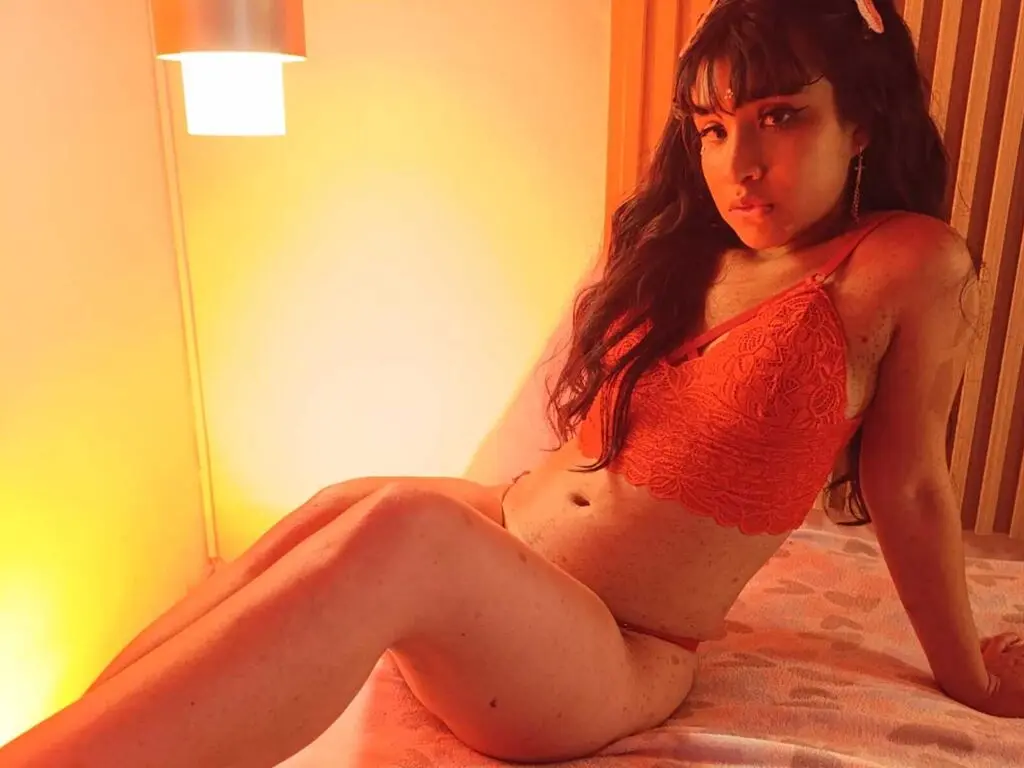 SharonRamoss wearing an orange lace bra and matching undies, sitting on a bed with long dark hair and bangs.