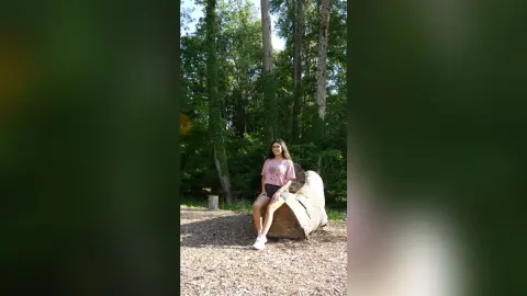 Young woman in a pink t-shirt sitting on a fallen log in a lush, sunny forest setting.