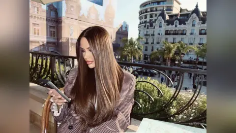 Brunette woman in a patterned blazer using a smartphone on a sunny urban balcony overlooking city buildings.