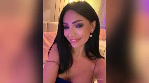 Smiling woman with long dark hair and glamorous makeup in a cozy, warm-lit bedroom setting.