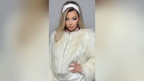 NicolePeach poses in a white furry coat and pearl necklace, wearing a white headband and heavy eye makeup against a light grey background.
