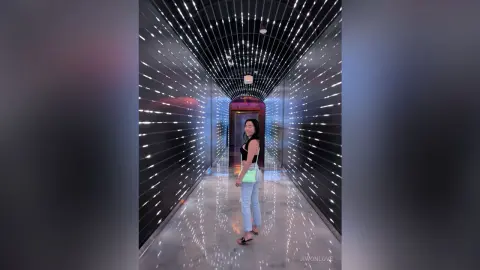 Smiling woman in a black tank top and jeans standing in a futuristic neon light tunnel hallway.