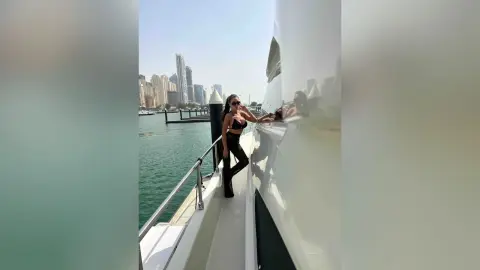 ArianaKroft stands on a white yacht deck wearing a black bikini top and black leggings against a backdrop of a city skyline and water.