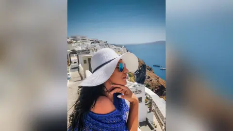 MissyJolie wears a white sun hat and a blue textured top while looking out over a coastal hillside view.