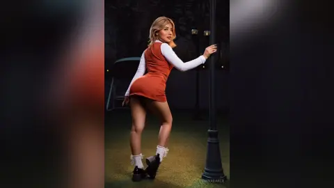 Blonde woman in a burnt orange mini dress and white long-sleeve shirt posing by a lamp post at night.