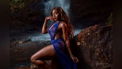 Elegant woman in a flowing blue sheer dress posing against a moody waterfall and dark rock backdrop.