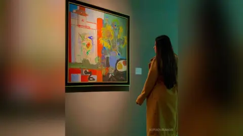 Woman in a long tan coat admiring colorful abstract floral artwork in a modern art gallery.