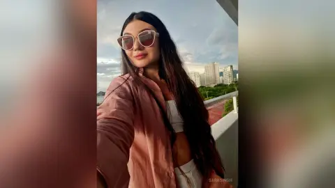 Smiling woman in pink shirt and sunglasses on a balcony overlooking city buildings at sunset.