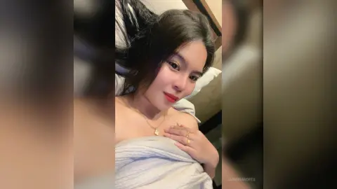 JasminAndres lies in bed with long dark hair, wearing a gold necklace and looking at the camera while wrapped in a white sheet.