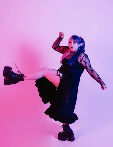 Gothic woman in a black lace dress and platform boots posing playfully against a pink studio background.
