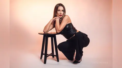 ChristineDavis poses in a black corset and black pants, leaning against a black stool against a plain light background.
