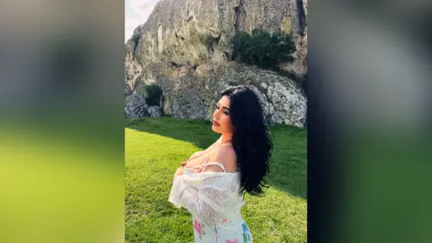 Beautiful woman with long black hair in a white lace top posing in a sunny green meadow by rocky cliffs.