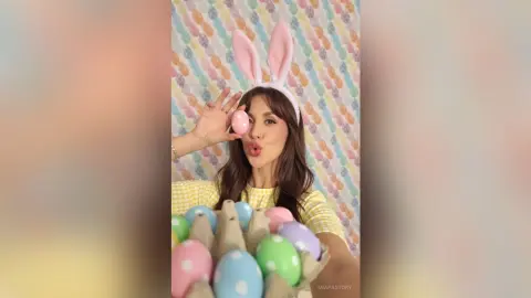 Cheerful woman in yellow top wearing bunny ears holding a pink Easter egg against a colorful egg pattern background.