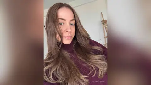 Young woman with long wavy brown hair wearing a cozy purple turtleneck sweater in a bright indoor setting.