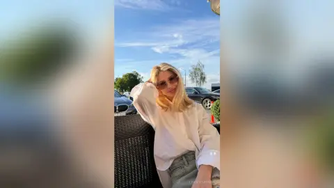 Stylish blonde woman in a white button-down shirt and jeans relaxing outdoors under a bright blue sky.