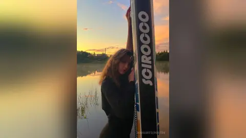 Young woman in black outfit leaning against a pole by a calm lake during a golden sunset mood.