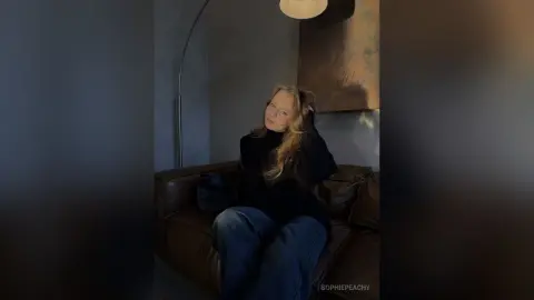 SophiePeachy sits on a brown leather couch wearing a black turtleneck and blue jeans.