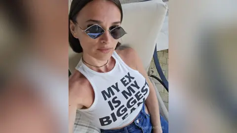 Woman in sunglasses and white graphic tank top relaxing outdoors in a sunny, casual mood.