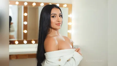AlessandraSanit poses with long black hair, wearing an off-the-shoulder white top in front of a lit vanity mirror.