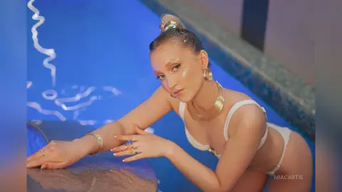 Glamorous woman in white bikini and gold jewelry posing by a glowing blue swimming pool at night.