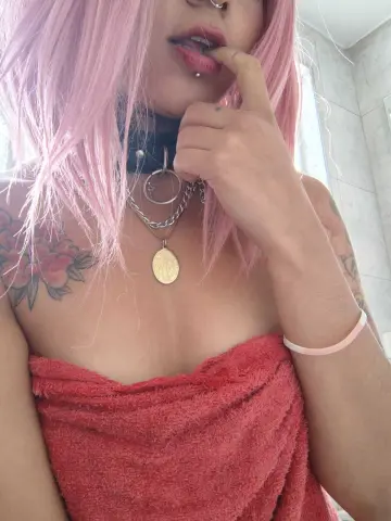 Woman with pink hair and floral tattoos wearing a red towel and silver layered necklaces in a bright setting.