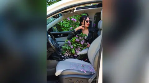 Stylish woman in a black leather jacket holding purple lilac flowers inside a luxury car.
