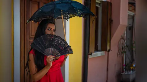 MissyJolie poses outdoors in a red dress, holding a black lace fan and a matching black lace parasol over her long dark hair.