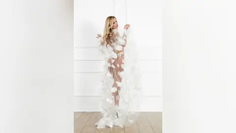 MilenaBia stands against a white wall wearing a sheer white floral lace bodysuit that shows her bare skin and curves.