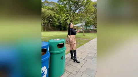Young woman in a black top and tan skirt exhaling smoke on a park path surrounded by lush green trees.