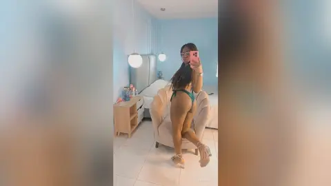 Young woman in a green bikini and clear heels posing in a bright, modern bedroom mirror selfie.