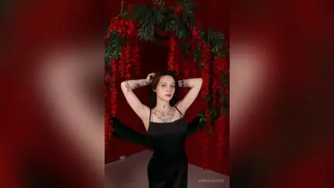 Tattooed woman in a black satin slip dress posing under a red floral archway in a moody, romantic setting.