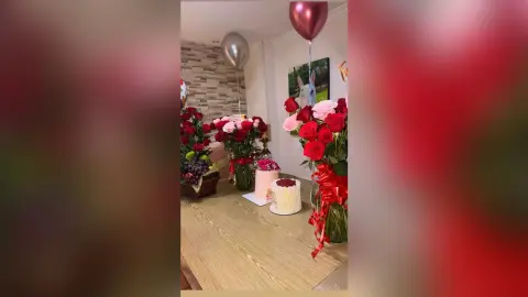 Elegant celebration setup with red rose bouquets, a white cake, and metallic balloons in a bright room.