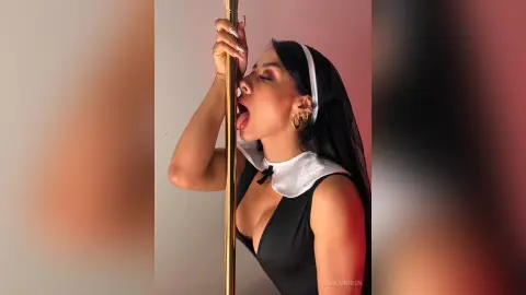 Glamorous woman in a black bodysuit and white collar posing playfully against a gold pole in soft lighting.