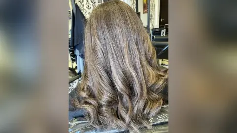Back view of long wavy ash brown hair with sun-kissed highlights in a salon setting.