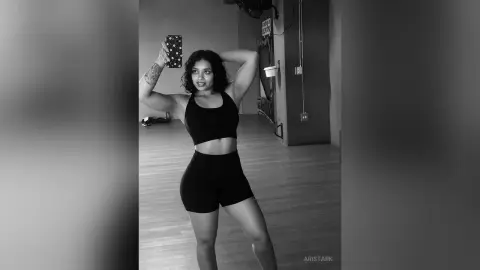 Confident woman in black athletic crop top and shorts posing for a mirror selfie in a minimalist studio.